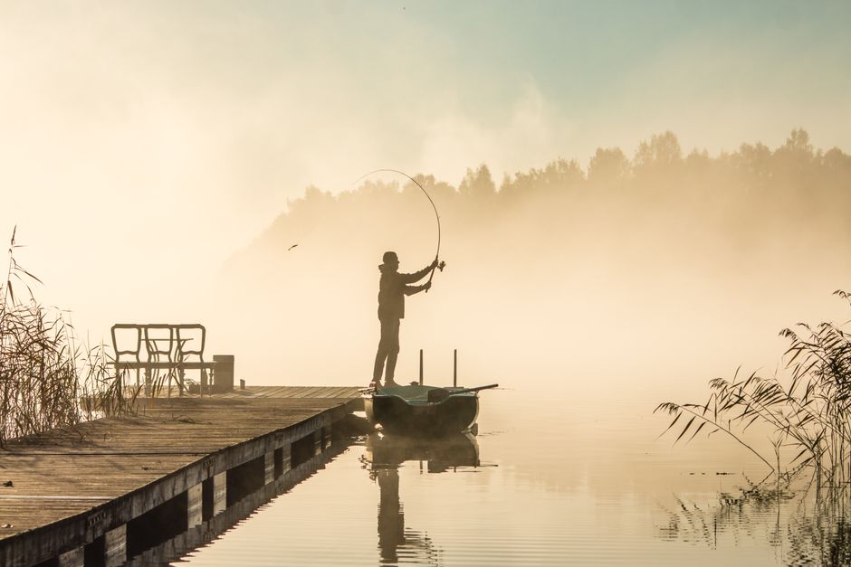 A tranquil scene of a lone fisherman casting a line on a misty lake at sunrise.