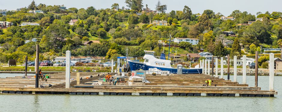 A boat dock under construction with workers in a scenic bay, surrounded by green hills and residential houses.