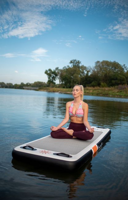 A woman practices yoga meditation on a floating mat in a tranquil lake setting.