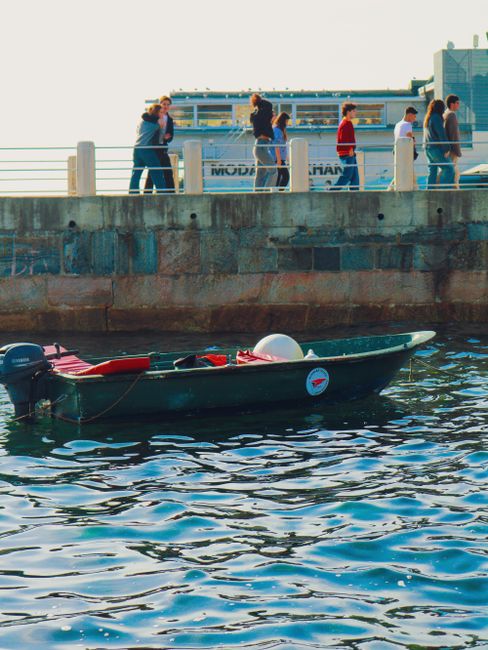 A small boat floating near a bustling urban harbor with people walking along the pier.